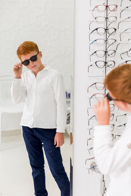 Photo gratuite garçon portant des lunettes noires et regardant dans le miroir au magasin d'optique