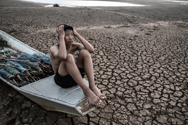 Le garçon s'est assis sur un bateau de pêche et a attrapé sa tête sur un sol sec, réchauffement climatique