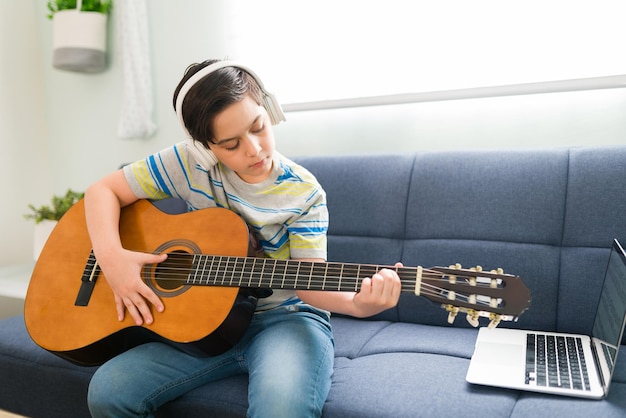 Garçon talentueux écoutant une chanson avec des écouteurs tout en jouant de la guitare acoustique et en suivant un didacticiel vidéo sur son ordinateur portable