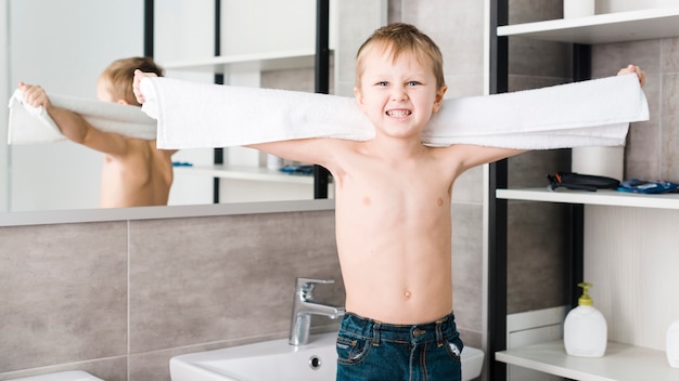 Un garçon torse nu tenant une serviette à la main en serrant les dents à la caméra dans la salle de bain