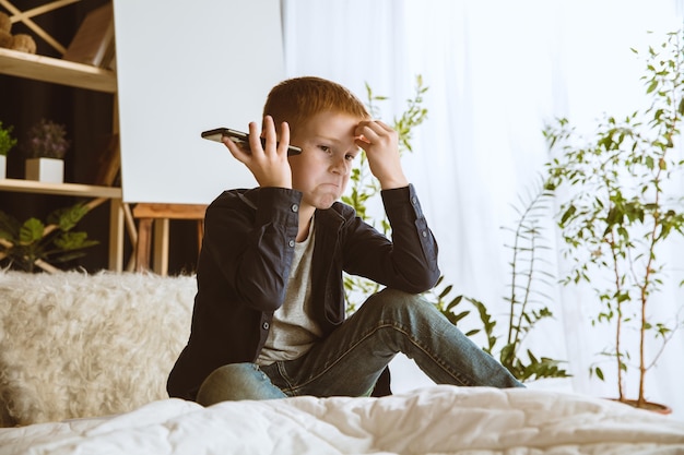 Garçon utilisant différents gadgets à la maison. Petit modèle avec montres intelligentes, smartphone ou tablette et écouteurs. Faire des selfies, bavarder, jouer, regarder des vidéos. Interaction des enfants et des technologies modernes.