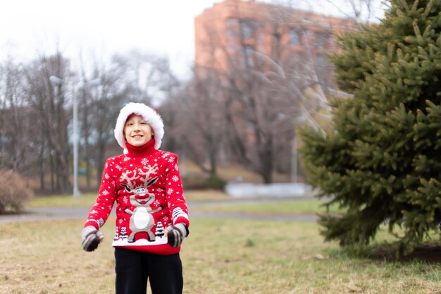 Un garçon vêtu d'un pull de noël chaud avec un cerf du nouvel an et un bonnet de noel s'est défendu et a caché ses mains dans le contexte d'un parc de la ville