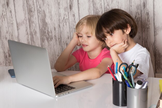 garçons mignons en t-shirts roses et blancs à l'aide d'un ordinateur portable gris sur la table avec des stylos sur fond gris