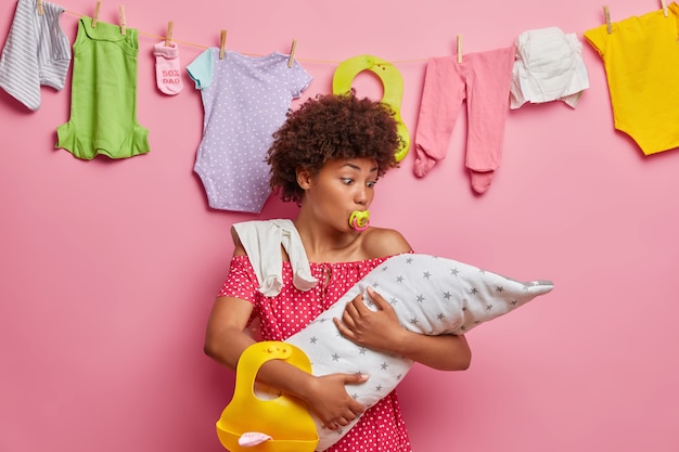 Photo gratuite garde d'enfants, concept de maternité. mère aux cheveux bouclés occupé embrasse le nouveau-né, pose avec des accessoires pour bébé, enfant qui allaite occupé