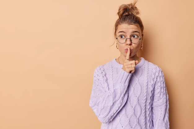 Gardez-le tranquille. Jeune femme appuie l'index sur les lèvres regarde mystérieusement loin fait un geste tabou fait signe chut le geste a besoin de silence potins sur quelque chose d'isolé sur le mur beige du studio