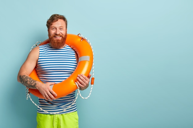 Photo gratuite un gars aux cheveux rouges barbu positif porte un gilet de marin rayé, porte une bouée de sauvetage orange