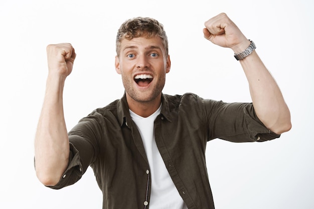 Photo gratuite un gars blond charismatique optimiste et excité aux yeux bleus et un large sourire blanc levant les poings serrés en signe de triomphe et d'encouragement souriant ravi du succès et de la victoire sur le mur gris