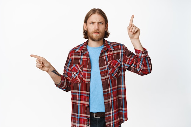 Photo gratuite un gars blond confus posant des questions et pointant sur le côté, pointant vers la gauche et vers le haut avec les sourcils froncés, voit quelque chose d'étrange, ne peut pas décider, perplexe debout sur fond blanc