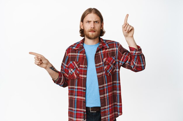 Un gars blond confus posant des questions et pointant sur le côté, pointant vers la gauche et vers le haut avec les sourcils froncés, voit quelque chose d'étrange, ne peut pas décider, perplexe debout sur fond blanc