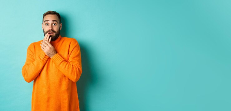 Photo gratuit un gars effrayé et choqué regardant la caméra debout dans un pull orange sur fond turquoise