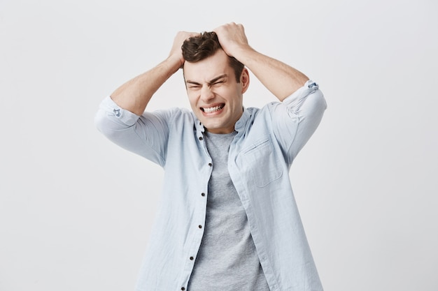 Un gars européen agacé et désespéré fronçant les sourcils, criant de colère, les yeux fermés arrachant les cheveux tout en se sentant furieux et fou de quelque chose. Expressions, émotions et sentiments négatifs sur le visage