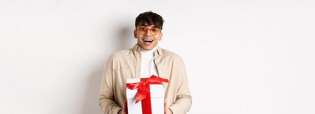 Un gars heureux surpris qui reçoit un cadeau tenant une boîte cadeau et souriant étonné debout sur une zone blanche