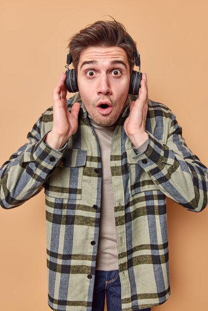 Un gars hipster impressionné regarde choqué par la caméra porte des écouteurs sans fil sur les oreilles écoute de la musique avec un son fort ne peut pas croire que ses yeux portent une chemise à carreaux décontractée isolée sur beige