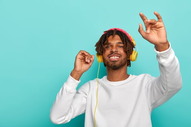 Un gars hipster à la peau sombre positive utilise des écouteurs modernes pour écouter de la musique, bénéficie d'un son parfait, sourit largement, danse contre le mur bleu