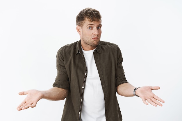 Photo gratuite un gars ignorant haussant les épaules comme n'étant pas au courant. portrait d'un homme blond mignon confus tenant la main sur le côté hébété, pinçant les lèvres et levant les sourcils interrogé contre le mur gris