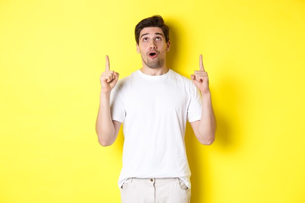 Gars impressionné vérifiant l'offre promotionnelle, regardant et pointant du doigt la publicité, debout sur fond jaune.