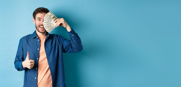 Photo gratuite un gars joyeux couvre la moitié du visage avec de l'argent et montre le pouce levé recommandant un prêt en espèces rapide permanent