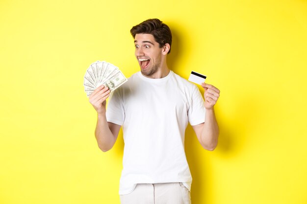 Un gars joyeux regardant de l'argent, tenant une carte de crédit, concept de crédit bancaire et de prêts, debout sur fond jaune