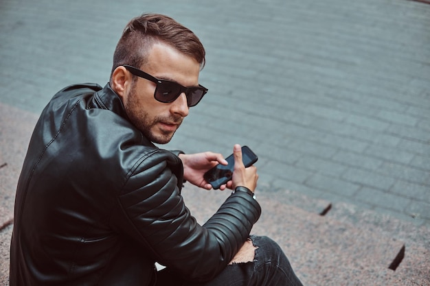 Un gars à la mode vêtu d'une veste noire et d'un jean assis sur des marches contre un vieux bâtiment en Europe.