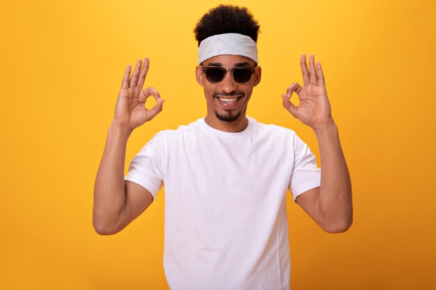 Un gars à la peau foncée en T-shirt blanc et des lunettes montre un signe ok sur fond orange Un homme gai en tee et un bandeau souriant largement sur isolé