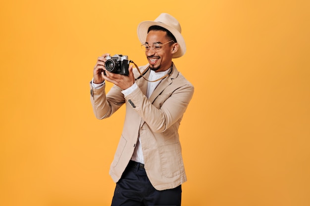 Photo gratuite un gars positif à lunettes et chapeau tient une caméra rétro et sourit sur un mur orange