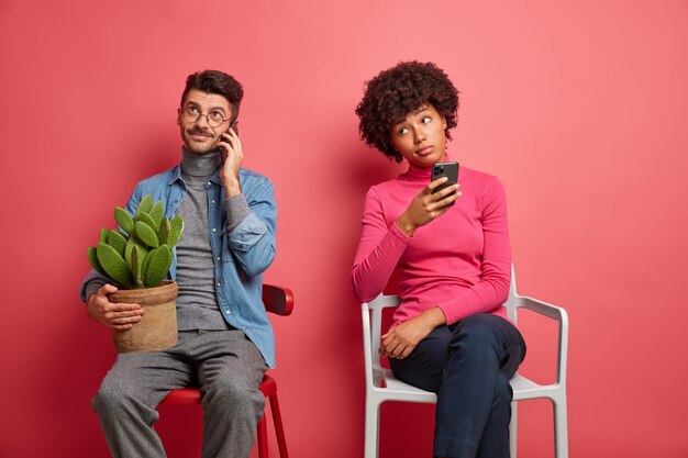 Un gars de race blanche a une conversation téléphonique tient un pot de cactus et pose à la maison sur une chaise. La femme ennuyée à la peau foncée tient cellulaire et réfléchit à la réponse à donner. Les gens et les technologies modernes