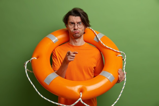 Photo gratuite un gars sérieux vous montre du doigt, pose avec une bouée de sauvetage gonflable, se soucie de la prévention des accidents, sourit narquoisement, porte des vêtements orange