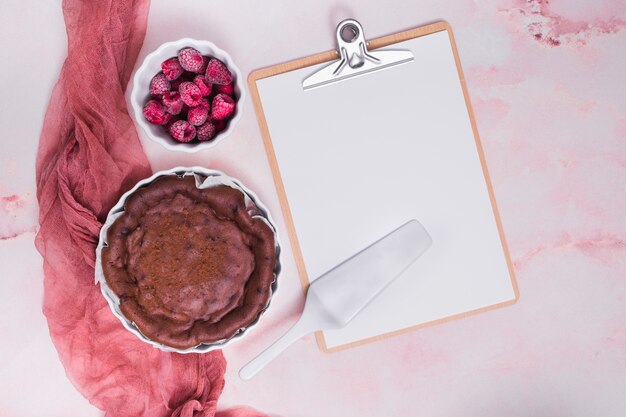 Gateau au four; framboise; spatule sur le presse-papiers avec du papier blanc sur le fond texturé rose