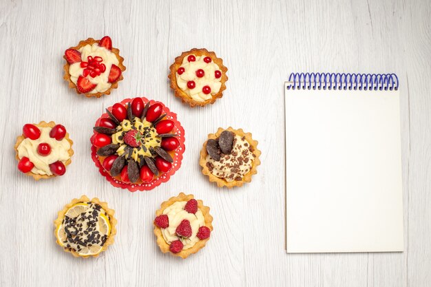 Gâteau aux baies vue de dessus sur les tartes de napperon en dentelle ovale rouge et un cahier sur le sol en bois blanc