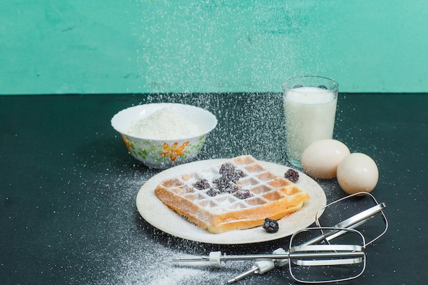 Photo gratuite gaufres en angle élevé dans une assiette avec des œufs, de la farine, du lait, des bâtons de mélangeur sur noir et cyan horizontal