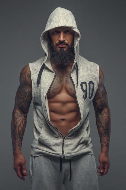 Gay tatoué musclé brutal en costume de sport gris avec capuche sur la tête. Isolé sur fond gris.