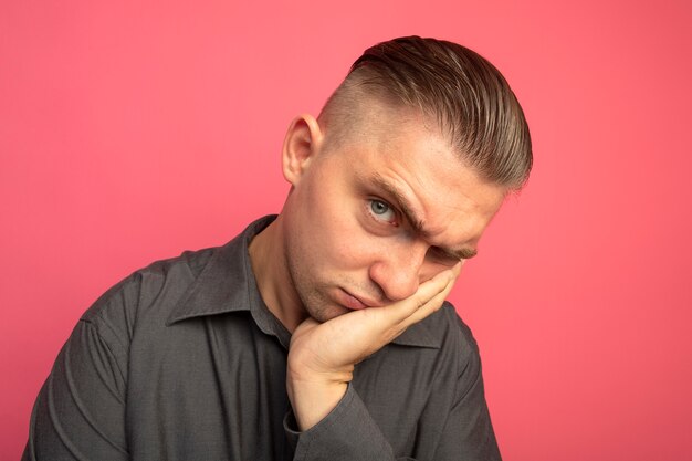 Gêné jeune bel homme en chemise grise regardant la tête penchée avant sur sa paume debout sur le mur rose