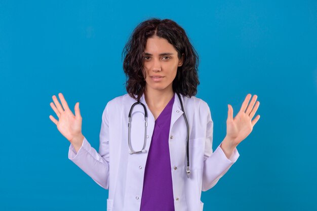 Gêné jeune femme médecin portant blouse blanche avec stéthoscope debout avec les mains levées sur bleu isolé