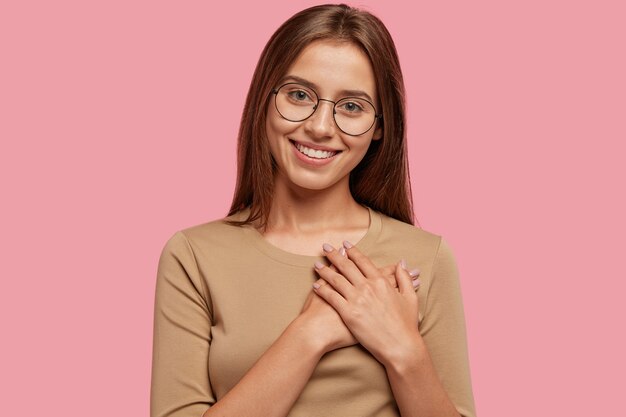 Généreuse fille à la recherche amicale reconnaissante garde les mains sur le cœur, a une bonne attitude, vêtue de vêtements décontractés