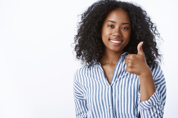 Génial bien fait. Amie à la peau foncée satisfaite et satisfaite portant un chemisier rayé souriant affirmatif montrant le pouce vers le haut comme approuver votre idée, souriant mur blanc satisfait