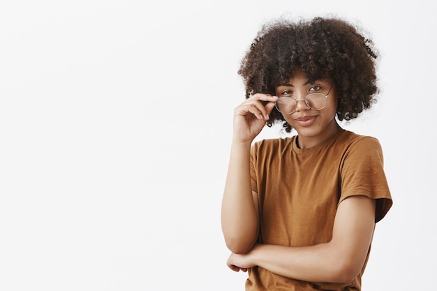 génie féminin afro-américain intelligent avec une coiffure afro dans un t-shirt marron à la mode à la recherche de sous des lunettes avec un regard sachant être méfiant souriant comme s'il avait un bon plan