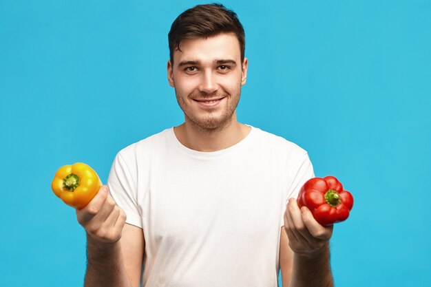 Les gens, les aliments biologiques, la nutrition, le végétarisme et le concept de mode de vie sain. Portrait de beau jeune homme positif portant un t-shirt blanc tenant des poivrons rouges et jaunes, va faire de la salade