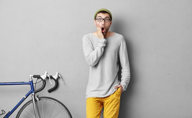 Gens, loisirs, activité, concept sportif. Jeune bel homme avec barbe, gardant une main sur la joue et l'autre dans la poche de pantalon jaune, choqué à cause du mauvais temps et des week-ends gâtés