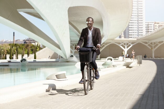 Les gens, le mode de vie, les transports et l'écologie. Beau gai souriant jeune employé de bureau à la peau sombre habillé officiellement pour se rendre au travail sur son vélo noir rétro, profitant d'un matin d'été ensoleillé