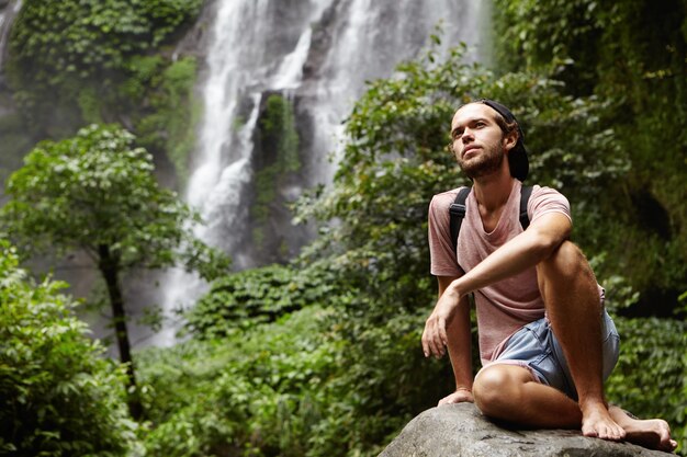 Les gens, la nature et l'aventure. Jeune hipster avec sac à dos assis sur un gros rocher en cascade