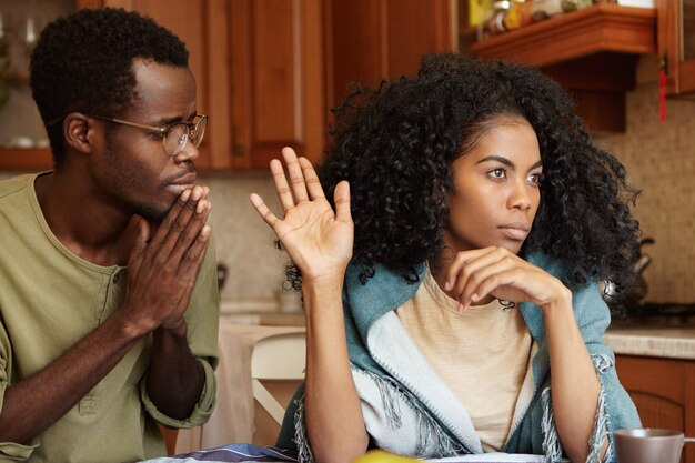 Les gens, les problèmes de relations et le divorce. Mâle repentant inquiet à la peau sombre gardant les mains pressées l'une contre l'autre, suppliant sa femme offensée de pardonner son infidélité, femme folle ne le regardant pas du tout