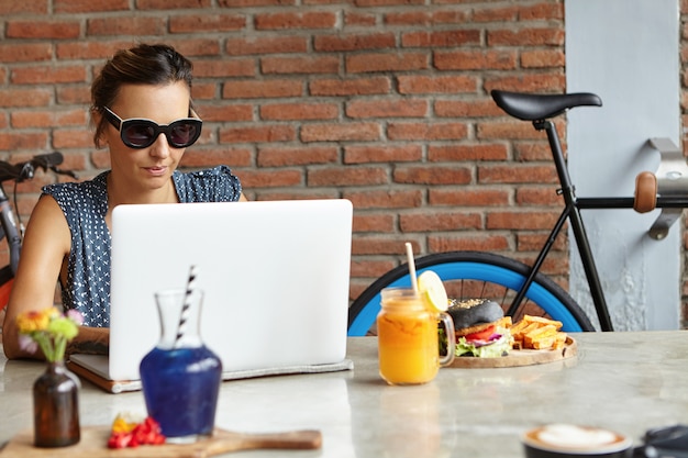 Les gens et la technologie. Femme d'affaires sérieuse et confiante habillée avec désinvolture à l'aide d'un ordinateur portable pour le travail à distance, assis à une cafétéria confortable avec mur de briques