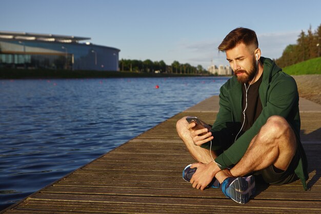 Les gens, les technologies modernes et le concept de communication. Vue extérieure du beau jeune hipster masculin mal rasé en baskets assis les jambes croisées en face du lac et écouter de la musique à l'aide de téléphone portable