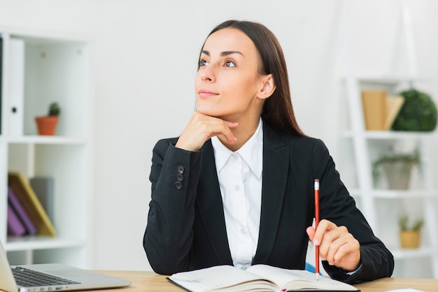 Photo gratuite gentil jeune femme d'affaires tenant un crayon sur son journal par-dessus le bureau