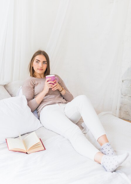 Gentil jeune femme tenant une tasse de café rose à la main, assis sur le lit