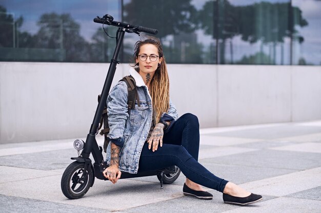 Une gentille fille pensive dans des verres et avec des tatouages sur son cou et ses mains se détend sur son propre scooter.