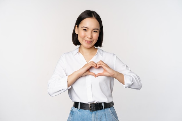 Geste d'amour Belle jeune femme asiatique montrant un geste du cœur et souriant exprimer des soins et de l'affection debout sur fond blanc