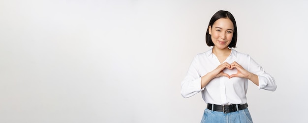 Geste d'amour Belle jeune femme asiatique montrant un geste du cœur et souriant exprimer des soins et de l'affection debout sur fond blanc