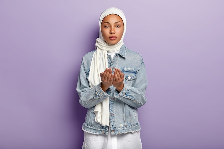 Photo gratuit geste de la main et concept de prière. une femme sérieuse à la peau sombre lève les mains en prière, plaide pour quelque chose, porte une écharpe et une veste en jean, isolée sur un mur violet. concept de religion musulmane
