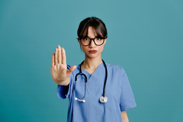 Geste montrant stricte jeune femme médecin portant un stéthoscope fith uniforme isolé sur fond bleu